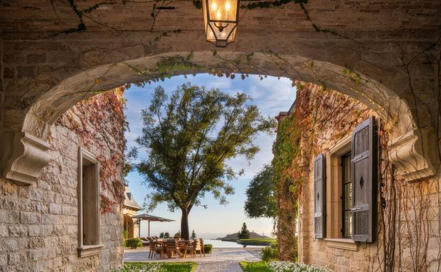 A historic stone and brick arched passageway opens onto a sunlit courtyard terrace with outdoor dining, a tree, clipped planting and a distant water view. Ivy-covered masonry walls, timber shutters and a hanging lantern create a rustic Mediterranean courtyard atmosphere.