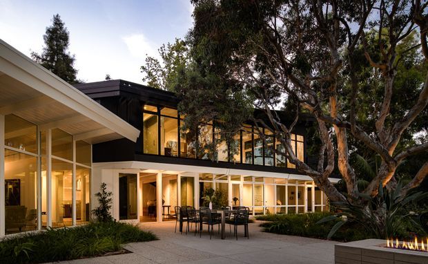Photograph of a contemporary, low-rise courtyard residence at dusk, with extensive floor-to-ceiling glazing, dark upper-level cladding, pale rendered walls, and broad overhanging roof planes. A paved outdoor dining terrace and linear fire feature occupy the foreground, while mature trees and planting frame the building.