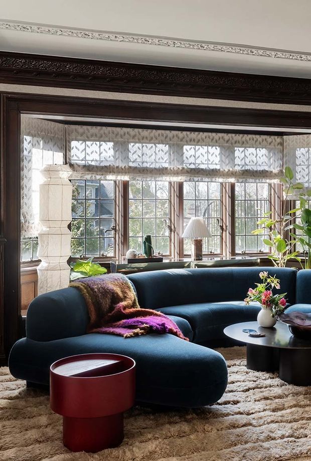 A richly styled living room viewed through a dark timber-framed opening, featuring a deep navy curved sectional sofa, textured neutral rug, sculptural red side table, black round coffee table, indoor plants, and a colourful throw. Tall multi-pane windows with patterned sheer curtains bring in abundant daylight. Ornate dark timber trim and decorative ceiling cornices add a traditional architectural character, while the furnishings create an eclectic, refined atmosphere.