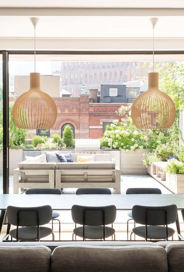 Bright, contemporary open-plan interior looking through full-height glazing onto a planted urban terrace. A long dining table with dark upholstered chairs occupies the foreground, with a pale upholstered sofa and outdoor-style seating beyond. Two large woven pendant lights hang from the ceiling, while raised planters filled with greenery frame views of surrounding brick buildings.