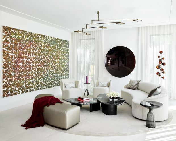 Bright, refined living room with white walls, sheer floor-to-ceiling curtains, a large abstract patterned feature artwork, sculptural cream seating, a matching ottoman, black round coffee tables, and a contemporary linear brass-toned ceiling light. A dark circular wall feature and decorative pendant sculpture add contrast, while a pale round rug unifies the seating arrangement.