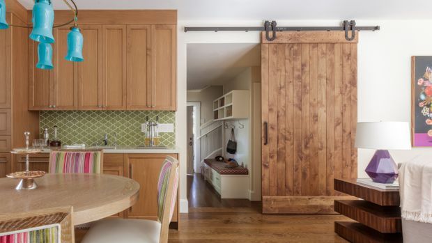Warm, eclectic open-plan dining and kitchen area with extensive natural timber cabinetry, a round timber dining table, patterned upholstered dining chairs, green geometric tiled splashback, and distinctive turquoise pendant lights. A rustic timber sliding barn door opens toward a hallway with built-in shelving and seating. The room combines traditional joinery with colourful contemporary furnishings and statement lighting.