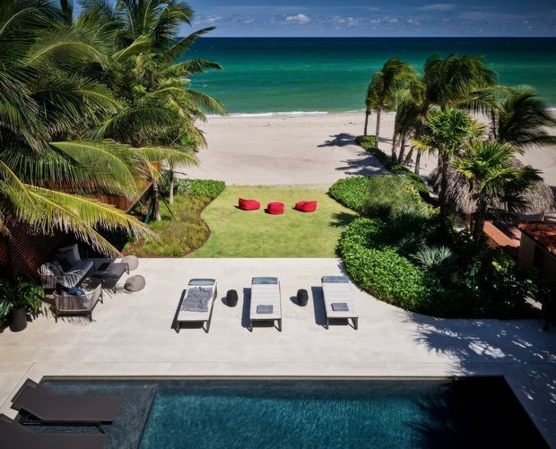 A tropical beachfront outdoor area viewed from an elevated position, featuring a rectangular swimming pool in the foreground, a pale paved sun terrace with loungers, a manicured lawn with red cushions, dense palm planting, and a sandy beach leading directly to turquoise ocean water.