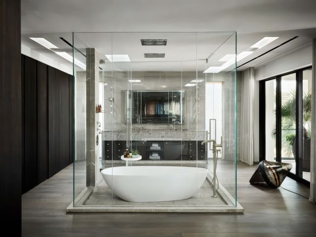 Contemporary bathroom suite featuring a freestanding white bathtub enclosed within a frameless glass shower enclosure. Dark timber or charcoal wall panelling and cabinetry contrast with a pale stone vanity and dark wood flooring. Large glazed doors, skylights, recessed ceiling lighting, and reflective surfaces create a refined, spacious feel. A separate toilet and vanity area is visible behind the glass enclosure.