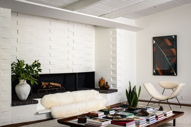 Bright, refined living room with a white-painted brick fireplace wall, exposed white-painted timber ceiling boards and beams, and a low dark hearth. A white sheepskin-covered sofa sits beside a mid-century-style lounge chair, while a low timber coffee table is covered with books and styling objects. Indoor plants and geometric wall art add restrained colour and texture.