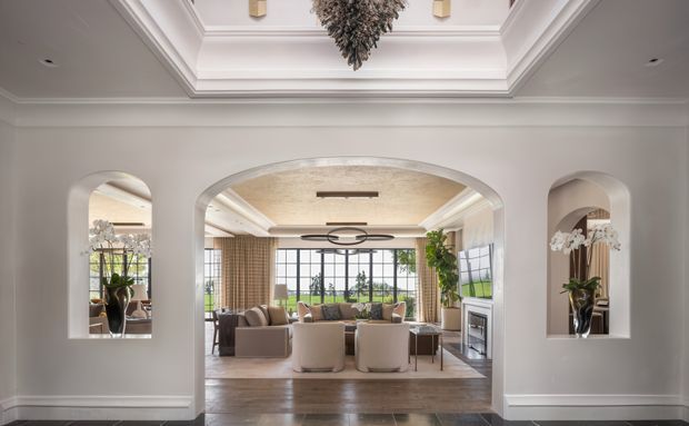 Elegant transitional living room viewed through a wide arched opening, with a coffered ceiling, white decorative wall mouldings, recessed arched niches containing orchids, warm timber flooring, and expansive black-framed glazing overlooking greenery. The room features a neutral seating arrangement, layered curtains, a fireplace, and refined contemporary lighting.