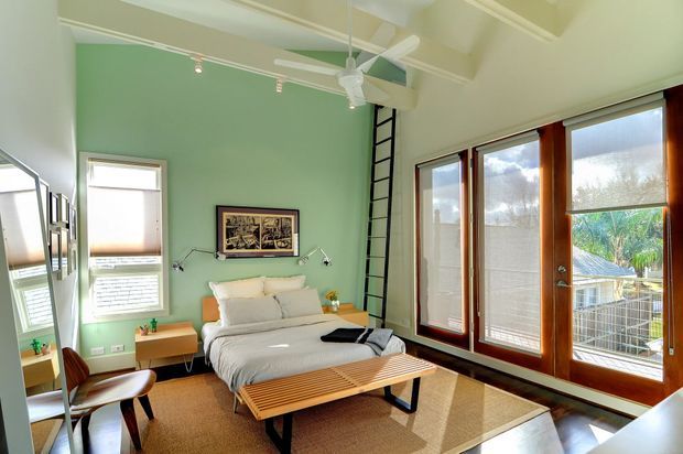 A bright, compact bedroom with a high angled ceiling and exposed white beams. The room features pale green walls, a low bed with neutral bedding, a timber slatted bench, a brown area rug, timber-framed glazed doors and windows, a ceiling fan, and a black ladder leading to an upper loft or storage area. A small desk and chair sit beside the bed, with a balcony or exterior view visible through the glazing.