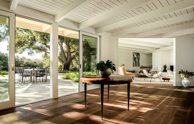Bright open-plan living and dining interior with expansive glazed doors opening to a landscaped outdoor terrace. The room features a white-painted exposed-beam and slatted ceiling, dark timber flooring, a round mid-century-style timber dining table with a potted plant, and a relaxed seating area beyond. Strong indoor-outdoor connection and abundant natural light create an airy, refined atmosphere.
