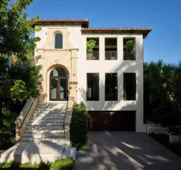 Front exterior view of a large two-storey stucco residence with a prominent Mediterranean-inspired entry pavilion, arched doorway and upper window, terracotta tiled roof edges, formal stair access, dark-framed windows, upper-level balcony openings, and an integrated garage. Dense tropical landscaping surrounds the home.