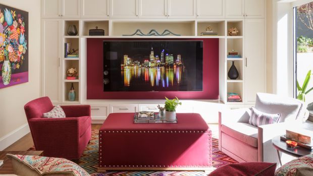 Bright, colourful living room centred on a large built-in white entertainment and display wall. A wall-mounted television sits within a deep burgundy-red feature niche, surrounded by white paneled cabinetry and open shelving. The room includes a burgundy upholstered armchair and ottoman, a white-and-pink armchair, patterned cushions and textiles, a round side table, and a large geometric patterned rug. Strong daylight enters from the right, creating a warm, inviting and eclectic atmosphere.