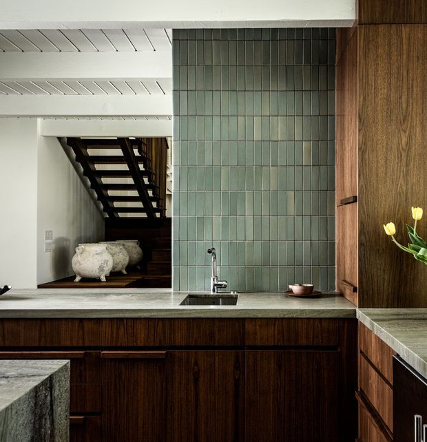 A sophisticated kitchen interior with dark wood cabinetry, a pale stone benchtop, and a muted green vertical tile splashback. A compact undermount sink and chrome tap sit beneath the tiled wall. The open-plan view reveals a dark timber staircase and sculptural vessels, while white painted timber ceiling boards contrast with the rich joinery.