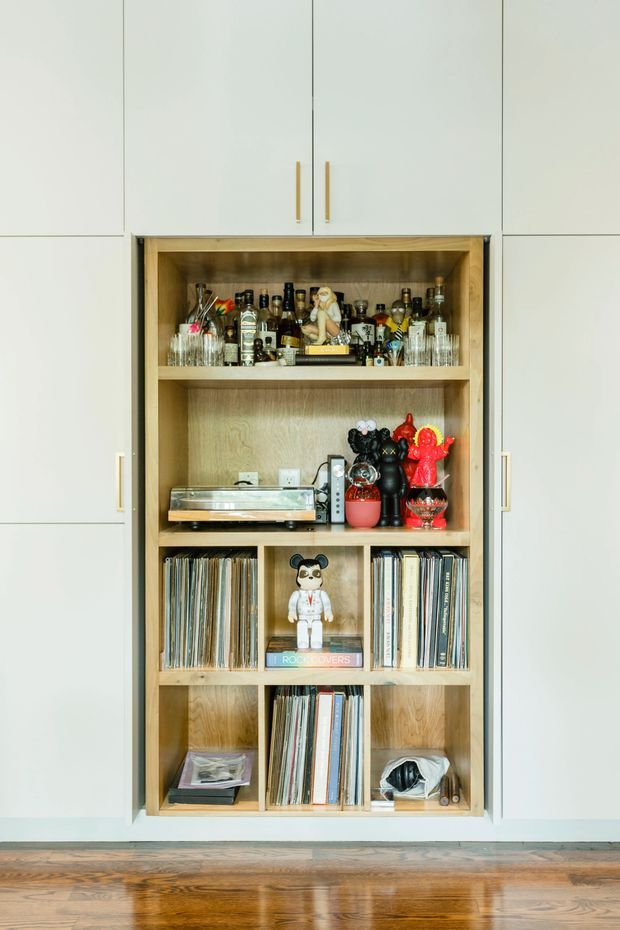 A built-in timber display and storage cabinet set into a white wall, with open shelves holding a home bar collection, glassware, a turntable, decorative objects, and vinyl records. The image also shows polished medium-toned timber flooring and minimal white cabinetry with slim brass-gold handles.