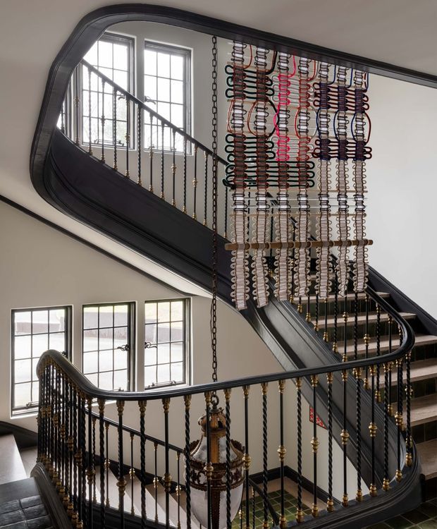 A grand interior stairway and landing with a sweeping curved staircase, dark painted timber handrails, ornate black-and-gold balusters, and a tall decorative chain installation hanging through the stairwell. Multi-pane windows provide abundant natural light, while the curved stair geometry and detailed metalwork create a dramatic heritage character.