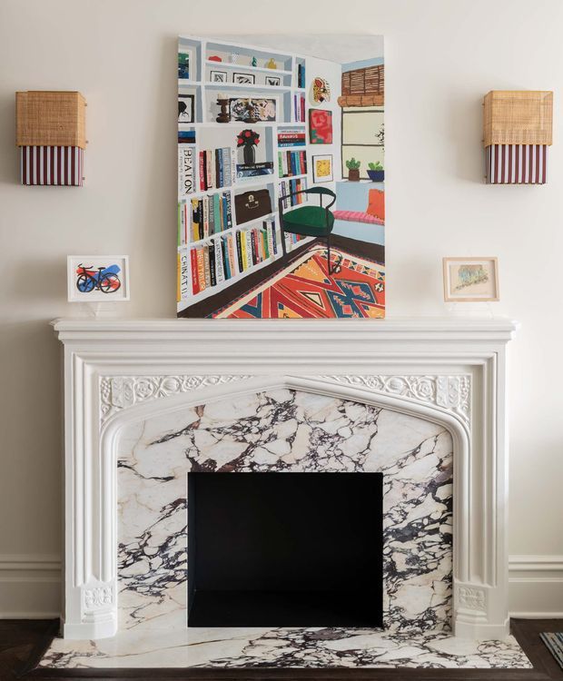 A refined interior detail centered on an ornate white fireplace mantel with carved classical mouldings and a dramatic white marble fire surround veined in dark grey and burgundy. A black firebox occupies the opening. Above the mantel is a colourful framed artwork depicting a densely arranged bookshelf and decor, flanked by two matching wall sconces with woven tan shades and red-and-white striped lower sections. Small framed artworks rest on the mantel, with neutral taupe walls and white trim completing the composition.