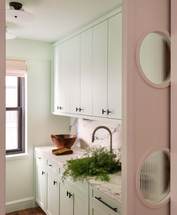 A compact, pastel-toned kitchen with pale mint-green walls and white cabinetry, featuring abundant upper and lower storage with slim black hardware. A marble-look stone benchtop and matching splashback surround a curved brushed-metal tap and integrated sink. A dark-framed window provides natural light, while a blush-pink partition wall with two round inset mirrors adds a distinctive decorative feature. Wood flooring and simple ceiling lights complete the space.