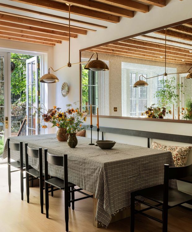 A warm, refined dining room with a long rectangular table covered in a small-check textile, surrounded by black dining chairs and built-in banquette seating. Exposed timber ceiling beams, pale walls, timber flooring, bronze pendant lights, and a large wall mirror create a bright, layered interior. Open glazed doors connect the room to a leafy exterior, while floral arrangements and patterned cushions add subtle colour.