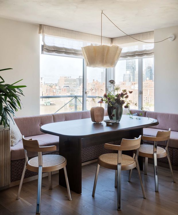 A bright high-rise dining nook with a large rounded rectangular dark wood dining table, built-in blush-toned banquette seating, and several mid-century inspired wooden chairs with woven cane seats and backs. Floor-to-ceiling corner glazing overlooks a dense city skyline, while a sculptural fabric pendant hangs above the table. Indoor plants, a floral arrangement, sheer Roman shades, and warm timber flooring complete the refined urban interior.