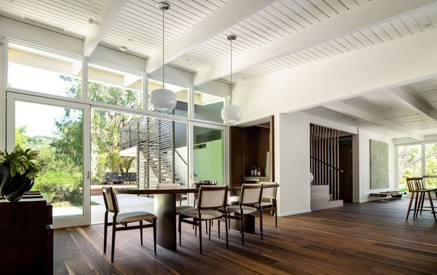 Bright open-plan residential interior featuring a dining area with a rectangular timber table and upholstered chairs, opening toward a living space and stair hall. Floor-to-ceiling glazing, white painted timber ceiling boards, dark timber flooring, timber screens, and sculptural pendant lights create a refined contemporary feel with strong indoor-outdoor connection.
