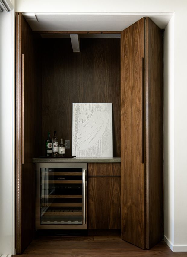 Compact built-in home bar or wine storage cabinet set into a wall niche, with dark walnut-toned wood cabinetry, a stone benchtop, integrated glass-front wine refrigerator, bottle storage, and concealed pocket-style doors. The cabinetry is framed by white painted walls and sits above a dark timber floor.