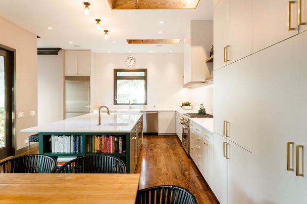 Bright contemporary kitchen viewed from the dining area, with a large central island, extensive white built-in cabinetry, timber flooring, warm timber ceiling inserts, and integrated open shelving filled with books. Black-framed glazing and a rear window bring natural light into the streamlined space. A timber dining table and black spindle-back chairs occupy the foreground.