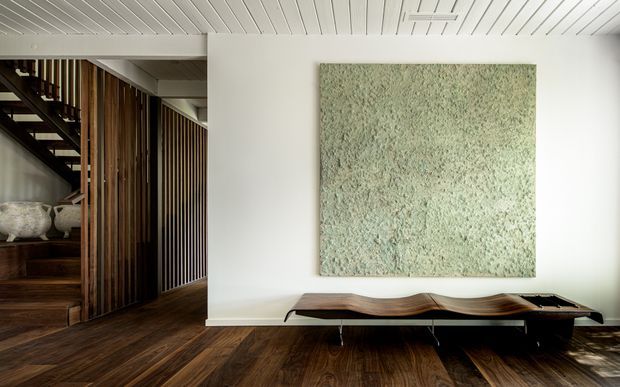A refined contemporary interior featuring a dark timber floor, white vertically panelled ceiling and walls, and a sculptural low timber console beneath a large textured pale green artwork. To the left, a timber staircase and vertical timber screen create a warm architectural transition into the adjacent space, with ceramic vessels displayed on open shelving.