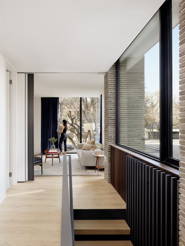 A bright, contemporary interior hallway with pale timber flooring, a short set of timber stairs, and a dramatic full-height black-framed window beside exposed light brickwork. A dark linear radiator sits above dark timber wall panelling beneath the glazing. The hallway opens toward a furnished living room with a cream sofa, dark curtains, a small round coffee table, and large windows overlooking trees. Clean white ceilings and walls contrast with the black and brick architectural elements.
