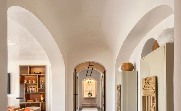 A warm, refined interior hallway with a long sequence of arched openings and a softly curved barrel-vaulted ceiling. Cream plaster walls and arches frame a central view toward a distant niche with a small plant. Built-in shelving on the left displays decorative objects and glassware, while tall pale cabinetry and woven wall decor line the right. Slim black hanging light fixtures and warm timber accents add contrast to the otherwise neutral scheme.