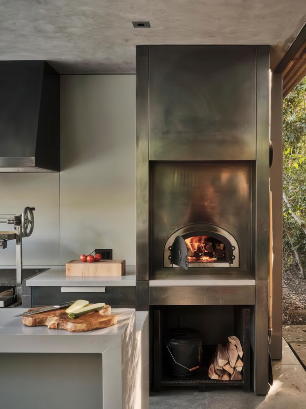 A compact contemporary kitchen centered on a tall dark metal built-in wood-fired oven and lower firewood storage. Sleek dark cabinetry and pale smooth wall and benchtop surfaces surround the oven, with a large island or workbench in the foreground. A timber chopping board, vegetables, and cooking equipment are visible, while a glazed opening at right looks onto trees and an exterior walkway.