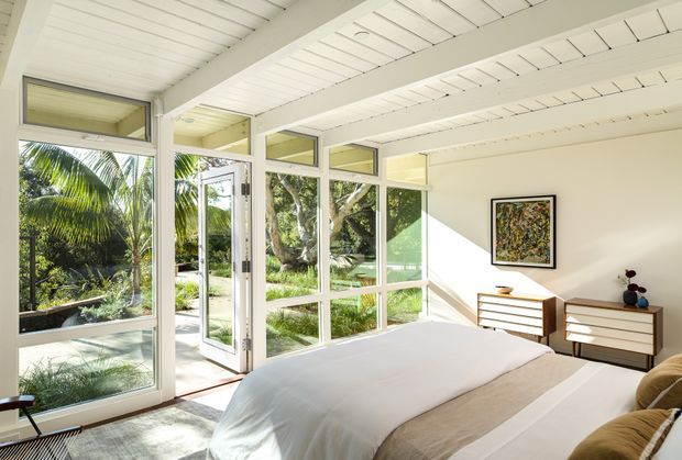 Bright, airy bedroom with a low white painted-beam ceiling, extensive floor-to-ceiling windows, and a glazed door opening to lush tropical landscaping. The room contains a large bed with neutral bedding, paired mid-century-inspired timber drawer units, and framed wall art. Strong indoor-outdoor connection and abundant natural light define the space.