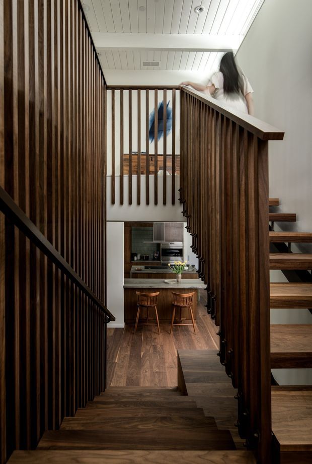 Interior view looking through a timber-lined stairwell toward a compact kitchen and breakfast counter. The space features dark stained timber stair treads, open-riser stairs, extensive vertical timber balustrade slats, a white painted timber-panel ceiling, and warm wood flooring. A person is visible on the upper landing, with a framed artwork and kitchen beyond.