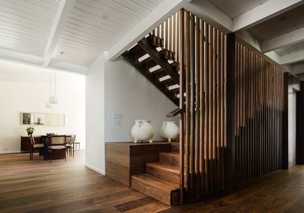 A warm, open-plan residential interior showing a timber staircase integrated with a dark wood slatted screen and built-in cabinetry. The stairs lead upward beside a dining area, while white painted structural beams and a white plank ceiling contrast with the rich timber flooring, stair treads, and vertical battens. The space has a refined contemporary feel with restrained furnishings and natural materials.