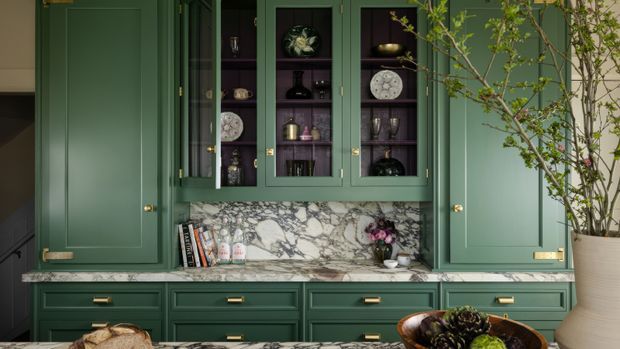 A richly detailed built-in green storage and display wall, featuring paneled cabinetry, glazed upper cupboards with decorative dishware and glassware, deep green drawers, and a dramatic grey-white veined stone benchtop and backsplash. A potted indoor tree and decorative bowl add organic accents.