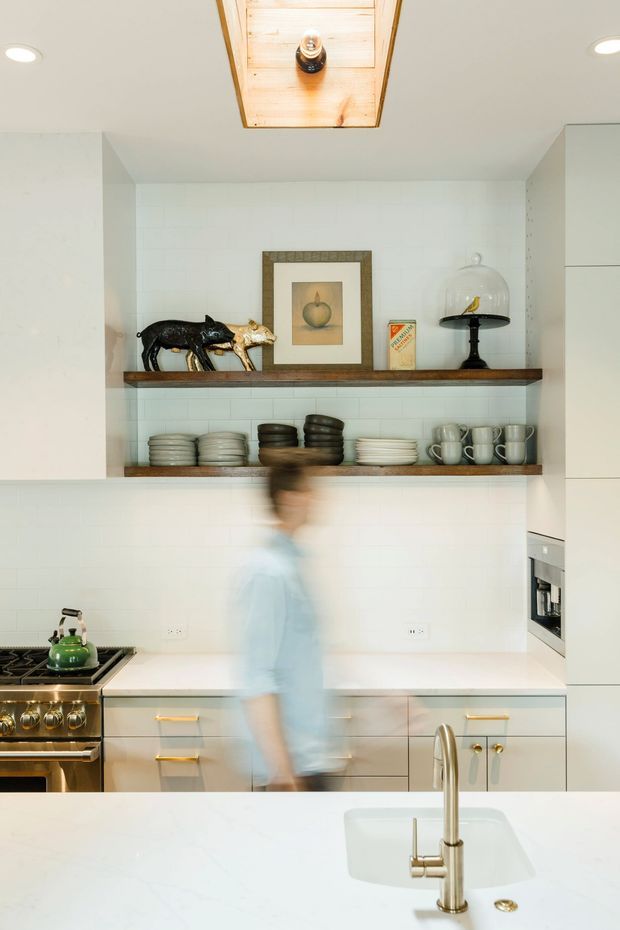 Bright, compact kitchen with white tiled walls, white cabinetry, a pale stone island benchtop and integrated stainless-steel appliances. Open timber display shelves hold crockery and decorative objects, while a timber-lined ceiling lightwell adds warmth. Brass-toned cabinet hardware and a vintage-style range introduce subtle character.