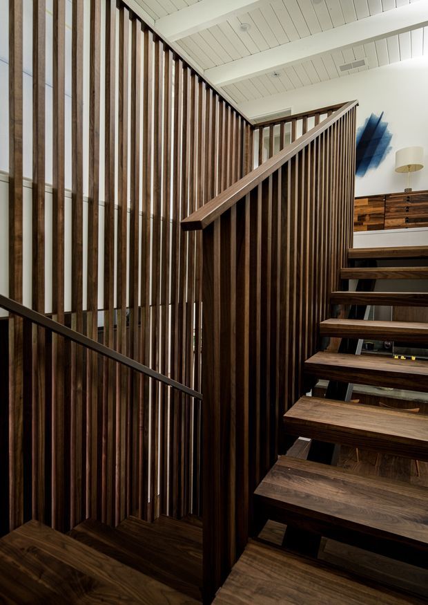 Interior architectural photograph of a contemporary open-riser staircase with dark timber treads, balustrade rails, and a dramatic vertical timber slat screen. The stair rises beside a white painted timber-lined ceiling and leads toward a landing with dark wood cabinetry and a blue abstract wall feature. Strong vertical lines, warm timber tones, and layered shadows dominate the composition.