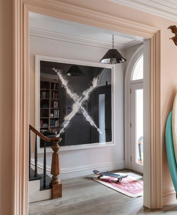 A refined traditional entry hall viewed through a wide, heavily moulded doorway. The space has pale painted walls, ornate cornices and wall panelling, a timber-and-black-metal staircase balustrade, pale timber flooring, a large framed monochrome artwork or mirror, and a pendant light. A tall arched-top glazed entry door brings in daylight. A skateboard and colourful rug add a casual contemporary touch, while surfboards are partially visible in the adjoining space.
