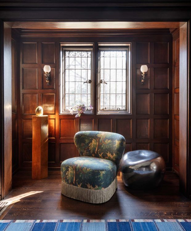 A richly detailed, dark wood-panelled interior with two leaded-glass windows, decorative wall sconces, a sculptural floral upholstered lounge chair, and a glossy rounded black side stool. Warm sunlight highlights the timber panelling and polished dark floor, while a blue patterned rug anchors the foreground. The space has a refined, historic character with eclectic contemporary furnishings.