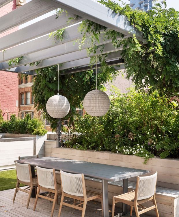 A landscaped urban outdoor dining terrace beneath a white-painted pergola, with lush climbing greenery, raised planter beds, a long dining table, woven timber chairs, and two spherical pendant lights. Brick and apartment buildings are visible in the background, creating a leafy courtyard atmosphere.