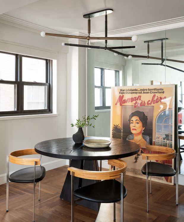 A bright dining area with a round dark timber dining table and curved timber-and-black chairs. The room has white walls, decorative ceiling moulding, dark-framed windows, a large mirrored wall, and a distinctive suspended black metal light fixture with exposed rods and globe bulbs. A vintage-style poster leans against the wall in the background. Herringbone timber flooring and restrained black, white, and warm wood tones create a refined contemporary interior.