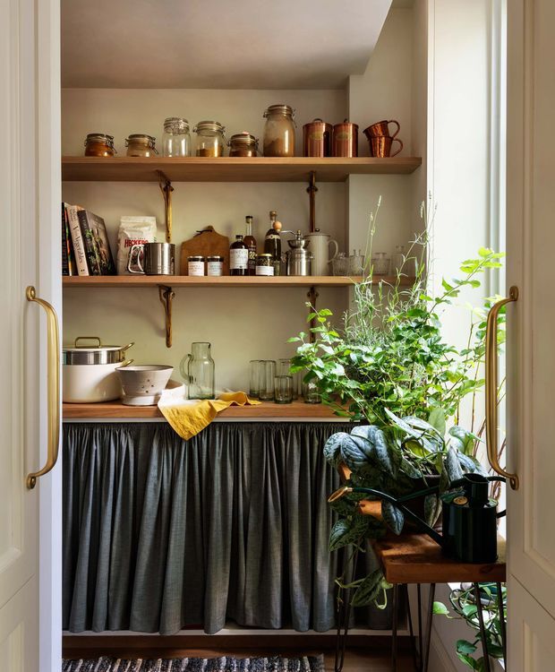 A warm, styled pantry or butler’s pantry viewed through open double doors. Open timber shelves display jars, cookware, bottles and crockery, with a lower counter concealed by a dark gathered fabric curtain. Brass-toned door hardware, a light neutral wall finish, and abundant leafy plants create an inviting vintage-inspired setting.