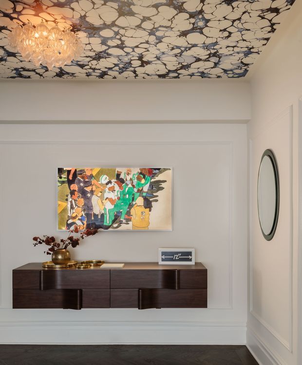 Elegant traditional-style entryway or hallway with white decorative wall panelling, a dark herringbone timber floor, and a floating dark wood console cabinet. A colourful framed artwork hangs above the console, accompanied by brass decor and a floral arrangement. The ceiling features a striking dark-and-cream organic patterned finish with a crystal chandelier, while an oval wall mirror is mounted on the right.