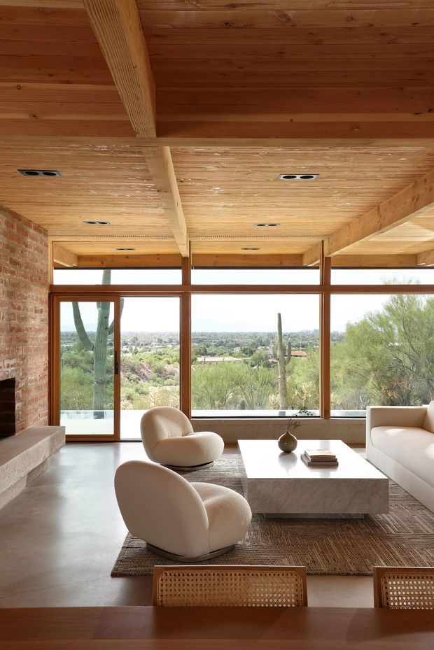 A warm, spacious living room with exposed timber ceiling boards and beams, exposed brick walls, expansive timber-framed glazing, and views toward a desert landscape. Cream sculptural lounge chairs and a sofa surround a large pale stone coffee table over a textured rug, with a dining table visible in the foreground. The room has a strong indoor-outdoor connection and abundant natural light.
