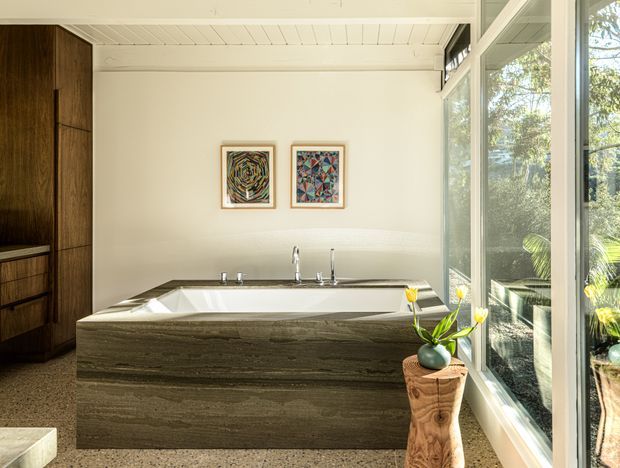 A bright, refined bathroom featuring a freestanding rectangular bathtub clad in horizontally layered dark stone, with wall-mounted and deck-mounted taps. A tall dark timber storage cabinet stands to the left, while full-height glazing on the right brings in garden views and abundant natural light. The white-painted timber-lined ceiling, terrazzo-like floor, framed artwork, and sculptural timber pedestal with flowers complete the space.