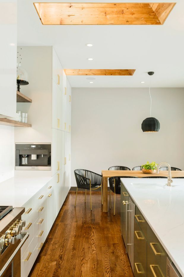 Bright contemporary kitchen and dining area with white full-height cabinetry, integrated stainless-steel appliances, a large white island with a raised gooseneck faucet, and warm dark timber flooring. The adjacent dining zone has a timber table, black woven chairs, and a dark faceted pendant light. Recessed ceiling lights and exposed warm timber ceiling panels add visual warmth and contrast.