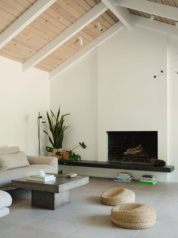 A bright minimalist living room with a tall white fireplace wall and a long dark hearth, furnished with a pale upholstered sofa, chunky concrete coffee table, woven round ottomans, books, and indoor plants. The room has a high pitched ceiling with exposed light timber boards and painted white structural rafters, large-format pale grey floor tiles, and understated contemporary lighting.