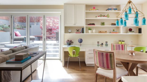 Bright open-plan dining and living area with a contemporary built-in white shelving and cabinetry wall, a round timber dining table with colourful upholstered chairs, and a dark rectangular living-room console in the foreground. Full-height glazed doors open to an outdoor area, while turquoise pendant lights and vivid accessories add playful colour. Timber flooring and neutral walls create a warm, airy setting.