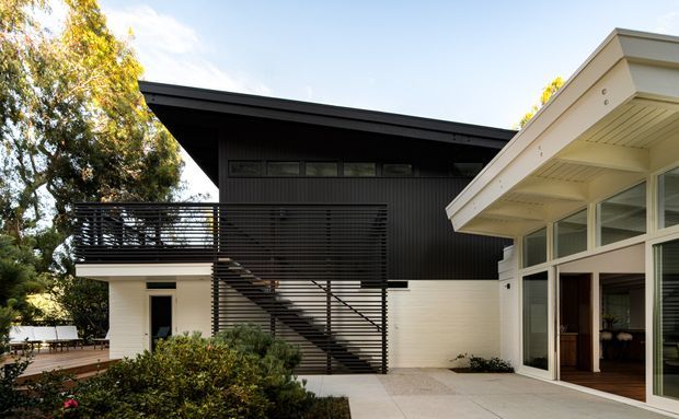 Exterior view of a contemporary multi-level residence with contrasting white masonry and dark vertical cladding. A prominent external steel staircase and horizontal slatted balcony screen define the facade, while expansive glazed doors open onto a paved terrace. Mature landscaping surrounds the building.