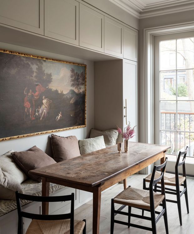 A refined dining area with a long, rustic wooden dining table surrounded by black ladder-back chairs with woven seats. Built-in pale grey cabinetry lines the wall and incorporates a cushioned banquette, while a large ornate framed artwork forms a feature above the seating. Tall traditional-style windows bring in abundant natural light, and layered crown moulding adds architectural detail.