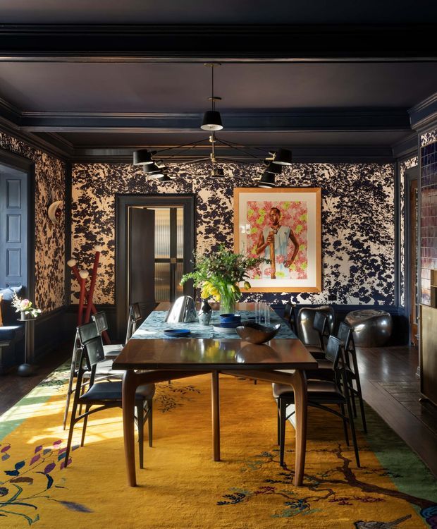 A richly decorated dining room with a long dark timber dining table surrounded by black leather and timber chairs. The room features dark blue-black panelled walls and ceiling with ornate patterned wallpaper, traditional cornice and trim detailing, a central pendant light, track-style artwork lighting, dark timber flooring, and a large vivid yellow patterned rug. A framed artwork and floral arrangement create colourful focal points.