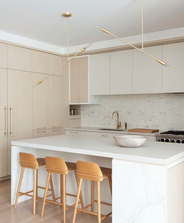 Bright, refined kitchen with a large white waterfall island and three timber bar stools. Floor-to-ceiling pale beige cabinetry lines the walls, paired with a light marble-look backsplash and matching island surface. Brass-toned hardware and sculptural linear pendant lights add warmth, while integrated appliances and a gas cooktop complete the contemporary fit-out.