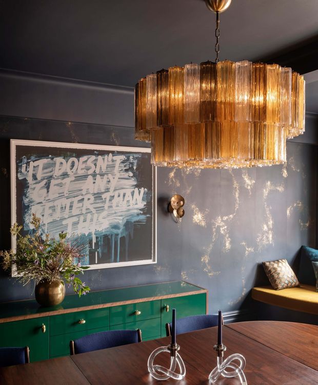 A moody, richly styled dining room with dark blue-grey textured walls, a large amber glass tiered chandelier, and a dark timber dining table with sculptural candleholders. A deep green built-in sideboard with brass hardware sits beneath an abstract artwork, while a window-seat bench with patterned cushions appears at right. The room has a glamorous, eclectic character with warm metallic lighting and layered decorative details.